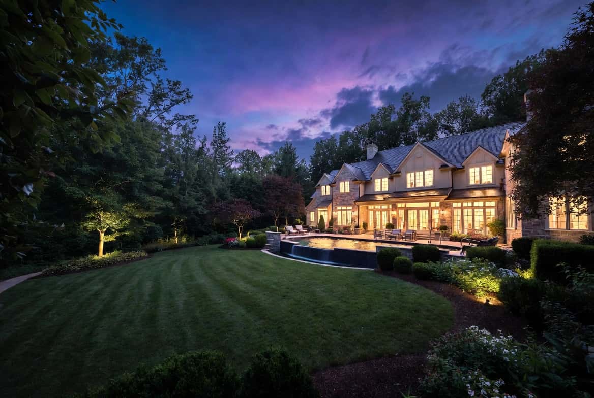 A stone-and-shingle luxury estate at twilight with warm window light, a lit pool, and a manicured lawn under a violet summer sky in North Fulton, Georgia